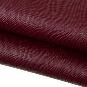 Könnte beinhalten: Nahaufnahme eines strukturierten, weinroten Ledermaterials. Das Leder hat ein schuppenartiges Muster und ist gefaltet, wodurch die Textur und Tiefe des Materials sichtbar werden. Die Farbe ist ein tiefer, satter Rotton.