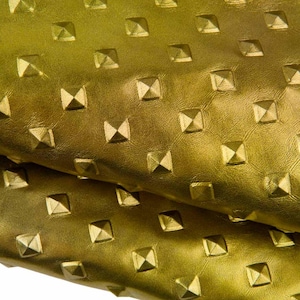 May include: A metallic gold-coloured material featuring a repeating pattern of raised, square-shaped pyramid studs. The surface has a smooth, leather-like texture with a consistent sheen, folded over to show depth and the uniform arrangement of the studs.