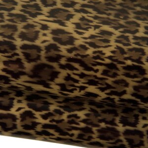 May include: A sheet of material featuring a repeating leopard print pattern in shades of dark brown, tan, and black. The surface has a smooth, glossy finish with a folded edge visible in the lower portion, showing the texture of the material.