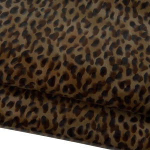 Pode incluir: Close-up de um tecido com padrão de leopardo. O padrão apresenta manchas castanhas escuras sobre um fundo bege. O tecido está dobrado, mostrando a textura e as camadas do material.