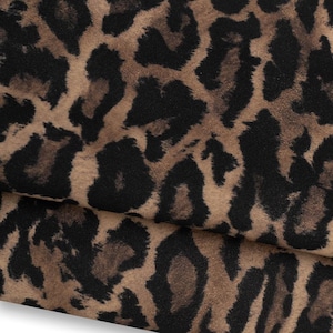 Könnte beinhalten: Ein gefaltetes Stück Stoff mit einem sich wiederholenden Leopardenmuster. Das Textil zeigt eine beigefarbene und braune Grundfarbe mit unregelmäßigen, tiefschwarzen Flecken und Markierungen, die in einem klassischen Tierfellmuster angeordnet sind.