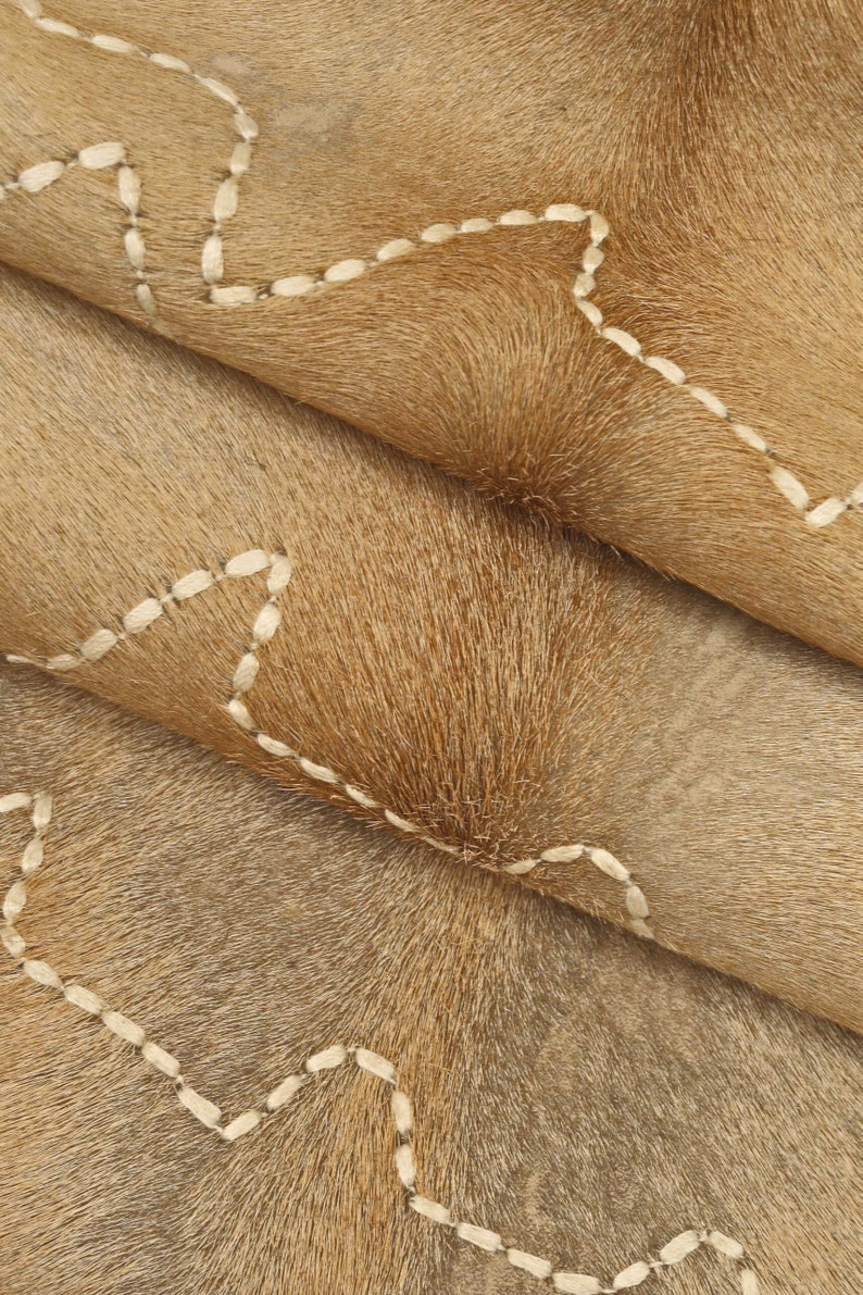 May include: Close-up of a brown cowhide rug with white stitching in a wavy pattern.