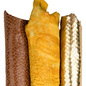 Może przedstawiać: Trzy rolki teksturowanej skóry w odcieniach brązu, żółci i złota. Brązowa skóra ma wzór łusek, żółta skóra jest pomarszczona, a złota skóra ma wzór rombów. Rolki są ustawione pionowo na białym tle.