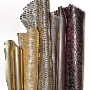 May include: A collection of metallic leather hides in various colors. The hides include gold, silver, and bronze tones, with some featuring a textured, reptile-like pattern. The leather is arranged vertically, showcasing the different textures and finishes.