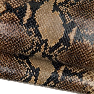 Peut inclure: Gros plan sur un motif de peau de serpent texturé. Le motif présente un mélange d'écailles marron, beige et noires, créant un design naturaliste et détaillé. Le matériau semble être du cuir.