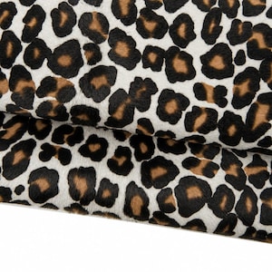 Puede incluir: Primer plano de una tela con estampado de leopardo. El patrón presenta manchas negras y marrones sobre un fondo blanco. La tela está doblada, revelando la textura y el patrón del material. Este textil es adecuado para diversos proyectos de artesanía y diseño.
