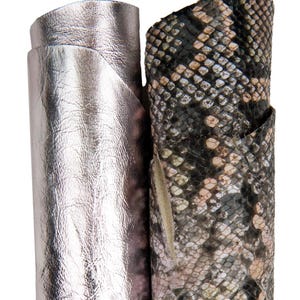 Op de afbeelding: Twee rollen leer. Eén is metallic zilverkleurig en de andere heeft een slangenpatroon in tinten zwart, bruin en beige. Het leer wordt waarschijnlijk gebruikt voor het maken van items zoals tassen, portemonnees of andere accessoires.