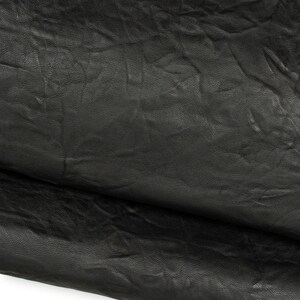 Può includere: Un pezzo di pelle nera con una consistenza distintamente rugosa e pieghettata. La superficie presenta una finitura opaca con pieghe profonde e motivi naturali della grana che creano un aspetto complesso e irregolare su tutto il materiale.