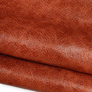 May include: A piece of textured, reddish-brown leather with a visible grain pattern. The material is folded over itself, creating a soft curve and a shadow along the crease, highlighting the uneven, rugged surface and the rich, warm color tones.