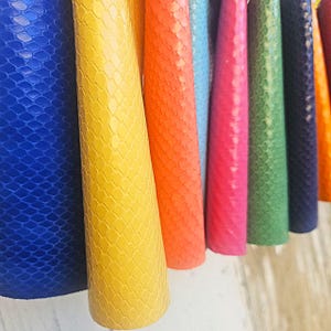 Op de afbeelding: Een close-up van verschillende rollen kleurrijk, getextureerd materiaal. De rollen zijn in tinten blauw, geel, oranje, lichtblauw, roze, groen en marine. Het materiaal heeft een schubbenachtig patroon.