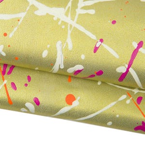 Op de afbeelding: Close-up van een getextureerd, goudgeel materiaal met witte, roze en oranje verfspatten. De verf is aangebracht in een willekeurig patroon, waardoor een levendig en artistiek ontwerp ontstaat. Het materiaal is gevouwen en onthult de textuur en kleurvariaties.