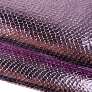 Peut inclure: Une feuille de matériau présentant un motif peau de serpent métallique violet et noir. La surface présente un design à écailles texturé et répétitif avec une finition brillante qui reflète la lumière, créant un aspect dimensionnel sur le tissu plié.