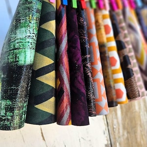 May include: A row of colorful fabric rolls, each held by a different colored clothespin. The fabrics display various patterns, including geometric shapes and abstract designs, in shades of green, yellow, purple, and orange.