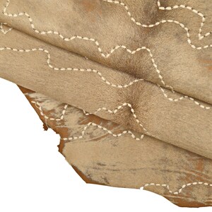 May include: A close-up of a light brown cowhide rug with white stitching in a wavy pattern. The rug is folded over itself, showing the texture of the hide.