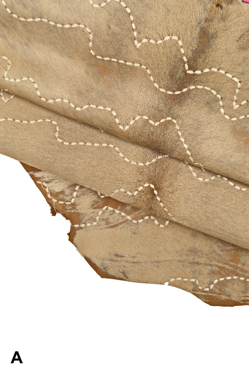 May include: A close-up of a light brown cowhide rug with white stitching in a wavy pattern. The rug is folded over itself, showing the texture of the hide.