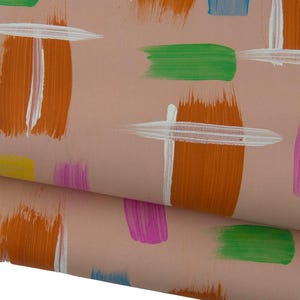Op de afbeelding: Een lichtroze stof met een herhalend patroon van geschilderde penseelstreken in levendige kleuren zoals oranje, felgroen, magenta, geel en blauw, waarbij sommige streken zijn bedekt met horizontale witte verfstrepen.
