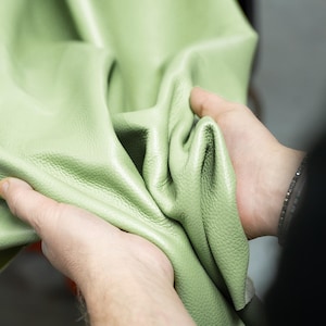 Könnte beinhalten: Nahaufnahme eines hellgrünen Lederstücks. Das Leder wird in zwei Händen gehalten, wodurch seine Textur und Flexibilität gezeigt werden. Das Material scheint weich und geschmeidig zu sein, mit einer subtilen Maserung.