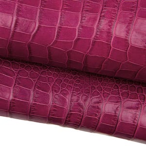 May include: Close-up of a roll of magenta faux crocodile leather. The material has a textured, embossed pattern that mimics crocodile scales. The leather is folded over, revealing the underside and the texture of the material.