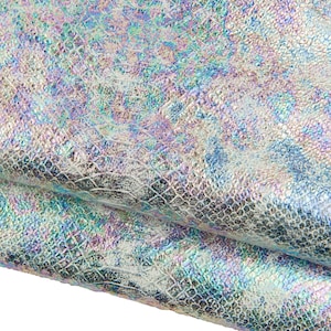 Peut inclure: Gros plan sur un tissu en similicuir irisé. Le matériau présente un motif texturé en forme d'écailles avec un effet chatoyant. Les couleurs incluent l'argent, le bleu, le vert et le violet. Le tissu est plié, révélant l'envers.