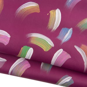 Op de afbeelding: Close-up van een diep magenta lederen materiaal met een herhaald patroon van kleurrijke penseelstreken. De penseelstreken zijn in tinten wit, geel, groen, blauw, roze en oranje. Het leer is in de linker benedenhoek omgevouwen.