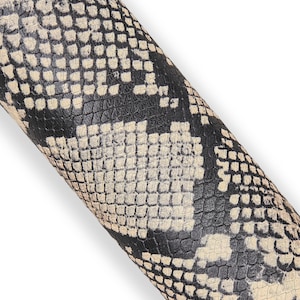 May include: Close-up of a rolled material with a snakeskin pattern. The pattern features a mix of black, cream, and off-white scales, creating a textured and detailed appearance. The material is likely leather or a leather-like product.