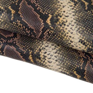 Puede incluir: Primer plano de un patrón de piel de serpiente texturizado. El patrón presenta una mezcla de escamas marrón oscuro, negras, beige y amarillas. El material parece estar doblado, revelando las variaciones de textura y color.