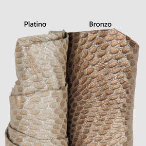 Puede incluir: Primer plano de dos piezas enrolladas de cuero texturizado. Una es de color plateado claro y metálico, etiquetada como "Platino". La otra es de color bronce más oscuro, etiquetada como "Bronzo". Ambas tienen un patrón similar a escamas.