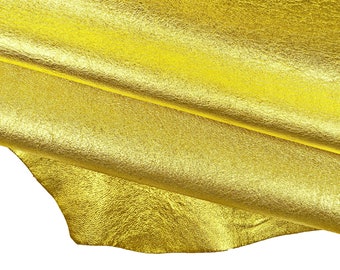 May include: A piece of metallic gold leather with a textured, crinkled surface. The material is folded over itself, creating soft curves and shadows that highlight the reflective, shimmering finish of the bright yellow-gold material.