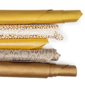May include: An overhead shot showcases several rolls of textured faux leather in various colors. The colors include gold, white with gold spots, yellow, snakeskin print, and a darker gold with a pebbled texture. The rolls are stacked on a white background.