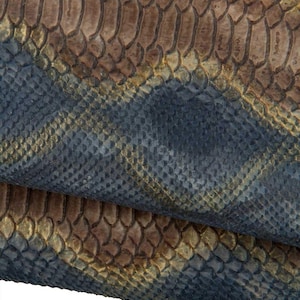 Op de afbeelding: Close-up van getextureerde slangenleer in tinten blauw, bruin en goud. De schubben zijn gedetailleerd en overlappen elkaar, waardoor een patroon ontstaat. Het materiaal lijkt gevouwen te zijn, waardoor de onderkant zichtbaar wordt.
