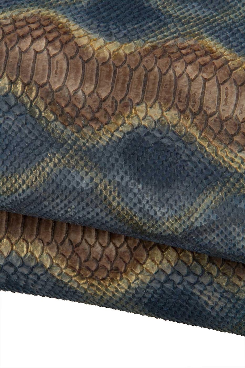 May include: Close-up of textured snakeskin in shades of blue, brown, and gold. The scales are detailed and overlapping, creating a pattern. The material appears to be folded, revealing the underside.