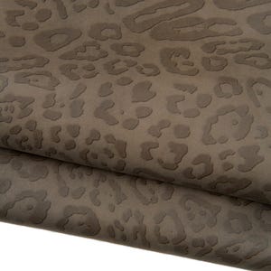 Puede incluir: Primer plano de una tela marrón doblada con un estampado de leopardo en relieve. La tela tiene un acabado mate y una paleta de colores neutros, adecuada para diversos proyectos de diseño. La textura es visible, con el estampado animal que añade interés visual.