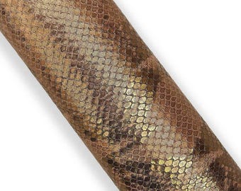May include: A rolled piece of faux leather with a snakeskin pattern. The material features a metallic gold and brown color scheme, with a textured, scale-like design. The material is angled diagonally across a white background.