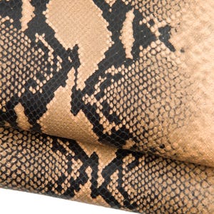 May include: Close-up of a snakeskin pattern. The pattern features a tan background with black and dark brown markings. The texture appears rough and scaly, with a natural, organic aesthetic. The image shows a folded piece of the material.