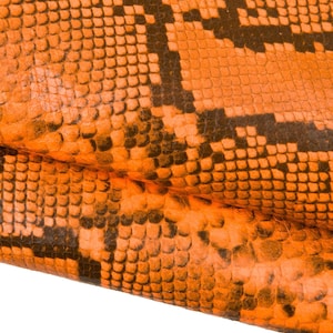 Può includere: Primo piano di un motivo a pelle di serpente arancione e nero. La pelle di serpente ha un aspetto strutturato e squamoso con un motivo ripetuto di forme arancioni e marrone scuro. L'immagine mostra una sezione piegata della pelle di serpente.