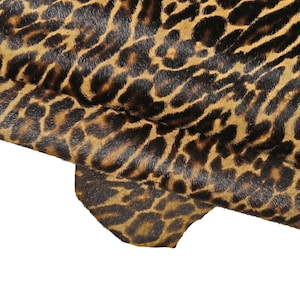 Puede incluir: Un trozo de piel animal con un estampado de leopardo con manchas de color marrón oscuro y negro sobre un fondo de color tostado dorado. El material tiene un acabado corto y texturizado similar al pelaje y está doblado para mostrar la letra C.