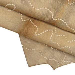 May include: A close-up of a light brown cowhide rug with white stitching in a wavy pattern. The rug is folded in half, showing the texture of the hide.