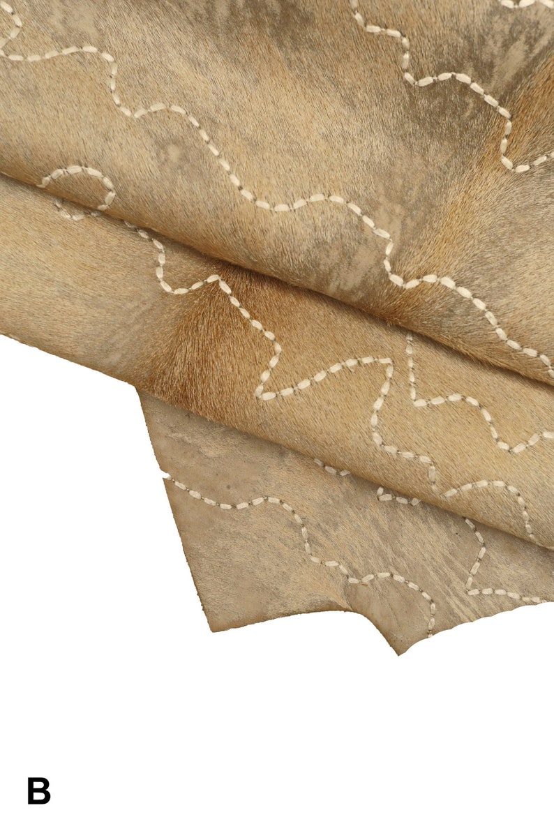 May include: A close-up of a light brown cowhide rug with white stitching in a wavy pattern. The rug is folded in half, showing the texture of the hide.