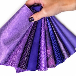 May include: A fan of various purple leather swatches featuring diverse textures including metallic, smooth, and embossed snakeskin patterns. The collection displays a range of violet and deep purple shades arranged in a layered, overlapping display.
