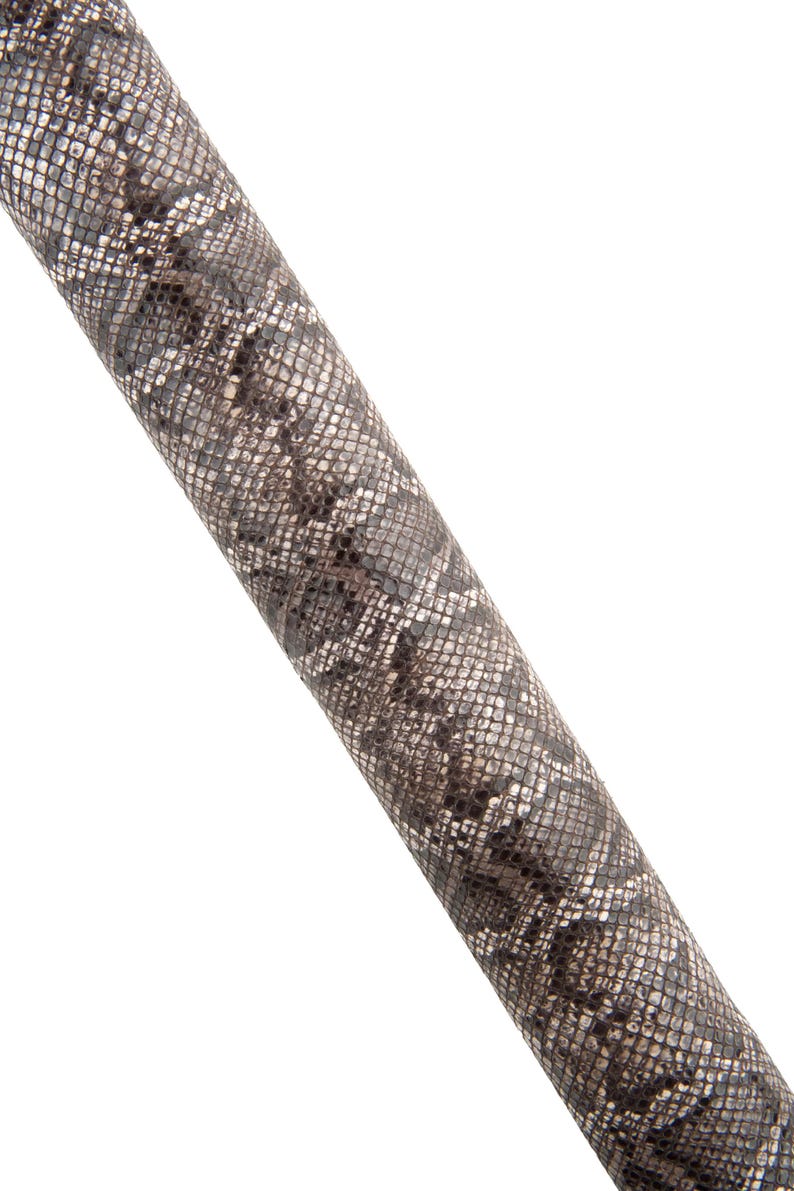 May include: Close-up of a cylindrical object with a snakeskin pattern. The pattern features a mix of grey, black, and white scales. The object is angled diagonally against a white background.