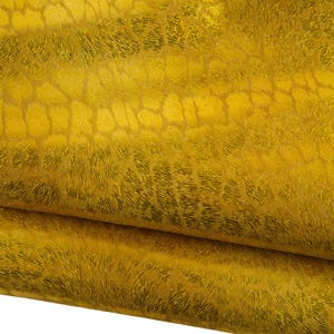 Könnte beinhalten: Nahaufnahme eines gefalteten Stücks goldgelben Leders mit einem strukturierten, reptilienartigen Muster. Das Leder hat eine glänzende, metallische Oberfläche und ein detailliertes, schuppenartiges Design. Die Falten zeigen die Dicke und Textur des Materials.