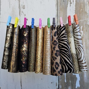 Puede incluir: Una colección de piezas de tela en forma de cono, cada una sujeta por una pinza de ropa de colores en una cuerda. Las telas muestran varios patrones, incluyendo lámina dorada, estampados de animales como leopardo y cebra, y diseños texturizados.