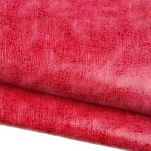 Puede incluir: Primer plano de un material de cuero rojo vibrante con un patrón texturizado similar al de un reptil. La superficie brillante refleja la luz, resaltando los intrincados detalles del diseño. El material está doblado, revelando su grosor y flexibilidad.