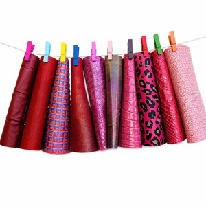 May include: Ten sheets of faux leather in various shades of red and pink, featuring textures like leopard print, crocodile skin, and geometric patterns, are hung on a string with colorful plastic clothespins against a plain white background.