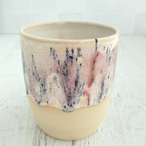 May include: A ceramic tumbler featuring a smooth, matte beige base and a glazed upper section. The glaze displays a blend of soft pink, cream, and white tones with vertical, dark blue-black drip patterns that create a textured, layered appearance.