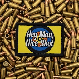 Könnte beinhalten: Ein rechteckiger Aufnäher mit dem Text "Hey Man, Nice Shot" in weißen Buchstaben, vor einem gelben Hintergrund mit einem blau-orangen Zielscheiben-Design. Der Aufnäher ist von zahlreichen messingfarbenen Patronen umgeben.