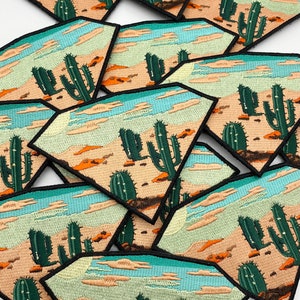 Puede incluir: Un montón de parches bordados para planchar que representan un paisaje desértico con cactus y un cielo azul. Los parches tienen forma de diamante con contornos negros.
