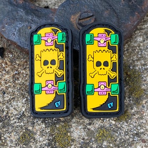 May include: Two black-bordered patches featuring a yellow skateboard design with a skull and crossbones graphic. The skateboards have green wheels, pink trucks, and the letters "EY" in blue. The patches are on a textured surface.