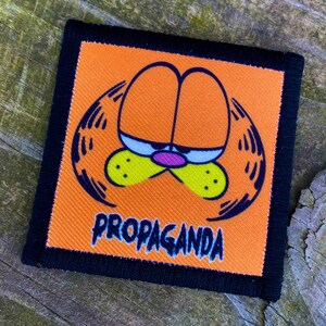 Può includere: Toppa ricamata arancione e nera con un viso di gatto dei cartoni animati e la parola "PROPAGANDA" in lettere bianche.