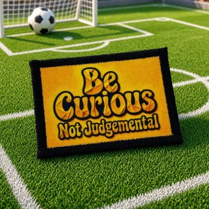 May include: A rectangular embroidered patch featuring a yellow background and a bold black border. The words "Be Curious Not Judgemental" are stitched in black with a textured yellow fill, positioned centrally on the patch, resting on a green football pitch.